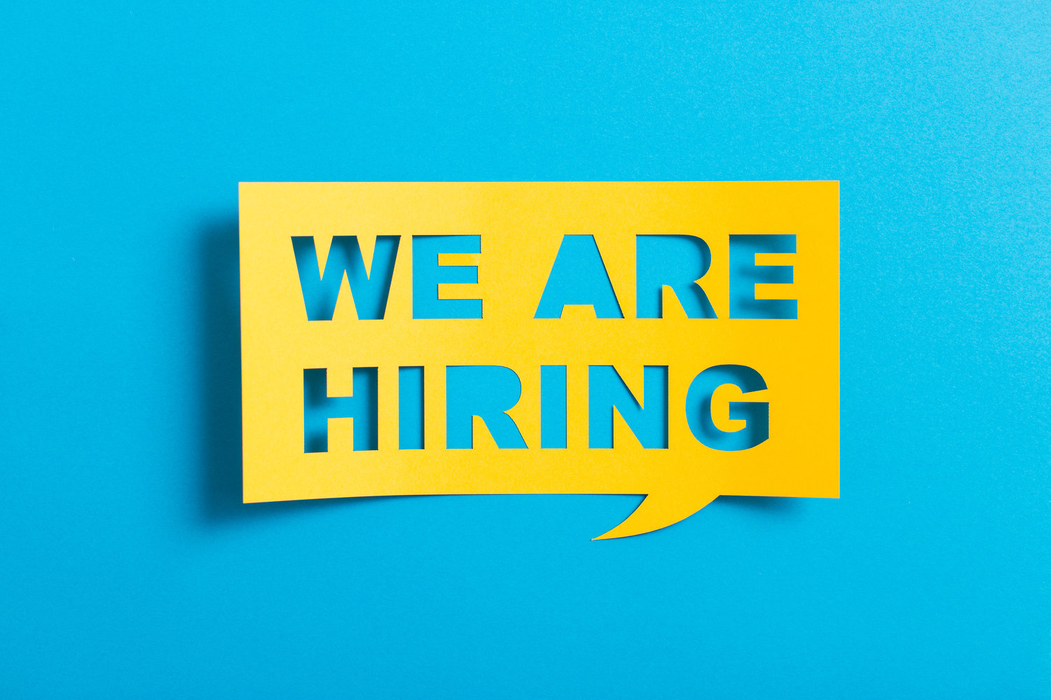 Een gele uitsnede met de tekst "WE ARE HIRING" op een blauwe achtergrond.