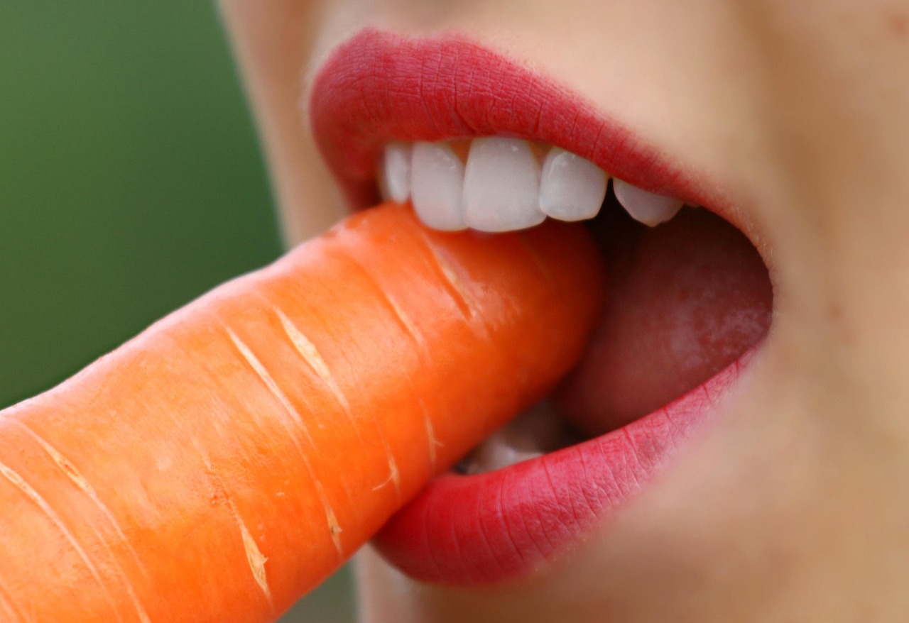 Een close-up van een glimlach met rode lippenstift en een wortel erin blazend.
